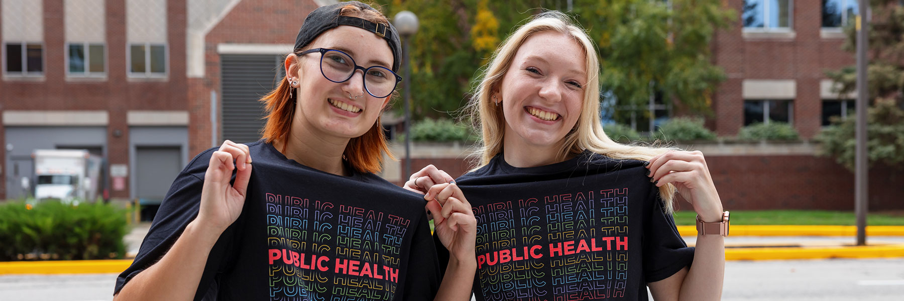 two smiling students showing off their public health t-shirts