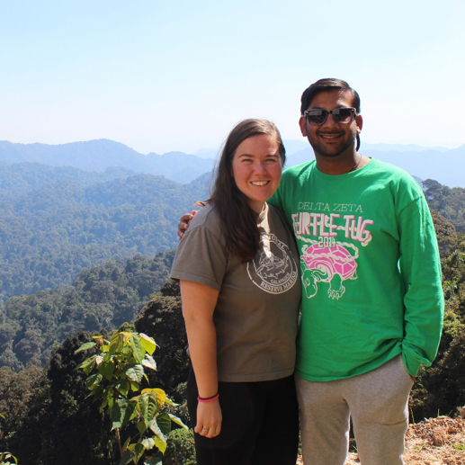 couple standing on cliff with mountains of trees in the background